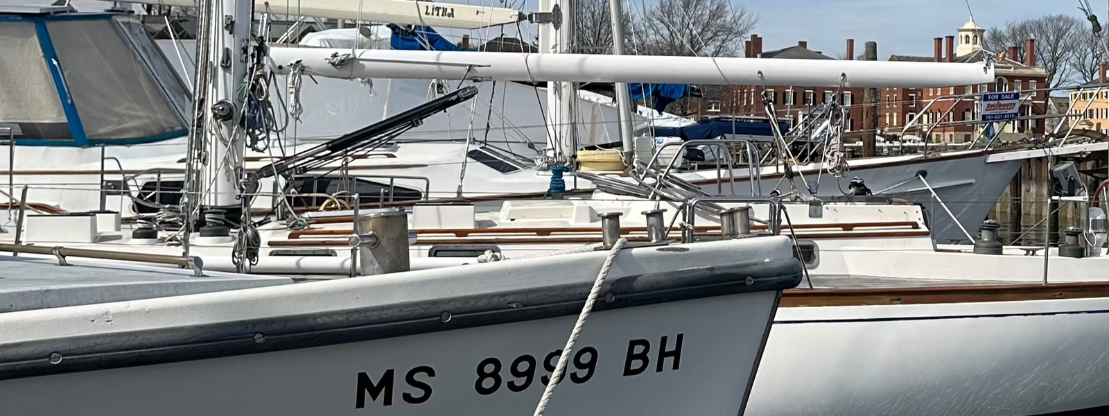 Close-up of a sailboat docked at a marina with bold black vinyl registration numbers “MS 8999 BH” on the hull, surrounded by other sailboats and dock infrastructure in the background.