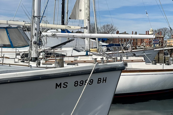 Close-up of a sailboat docked at a marina with bold black vinyl registration numbers “MS 8999 BH” on the hull, surrounded by other sailboats and dock infrastructure in the background.